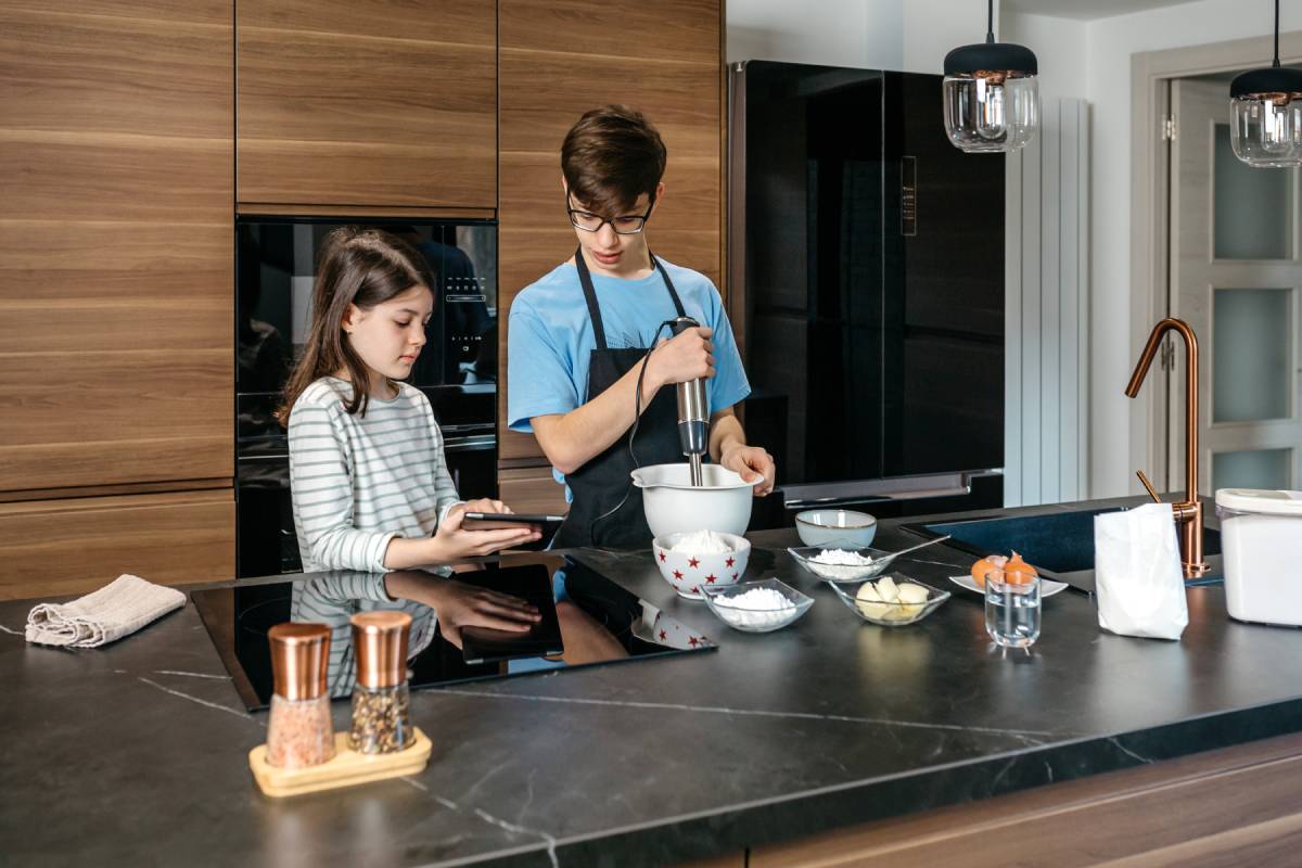 Two teenagers are making a cake in modern kitchen, one using hand mixer while other consults a recipe on electronic tablet. Brother and sister cooking together. Family leisure time at home concept.